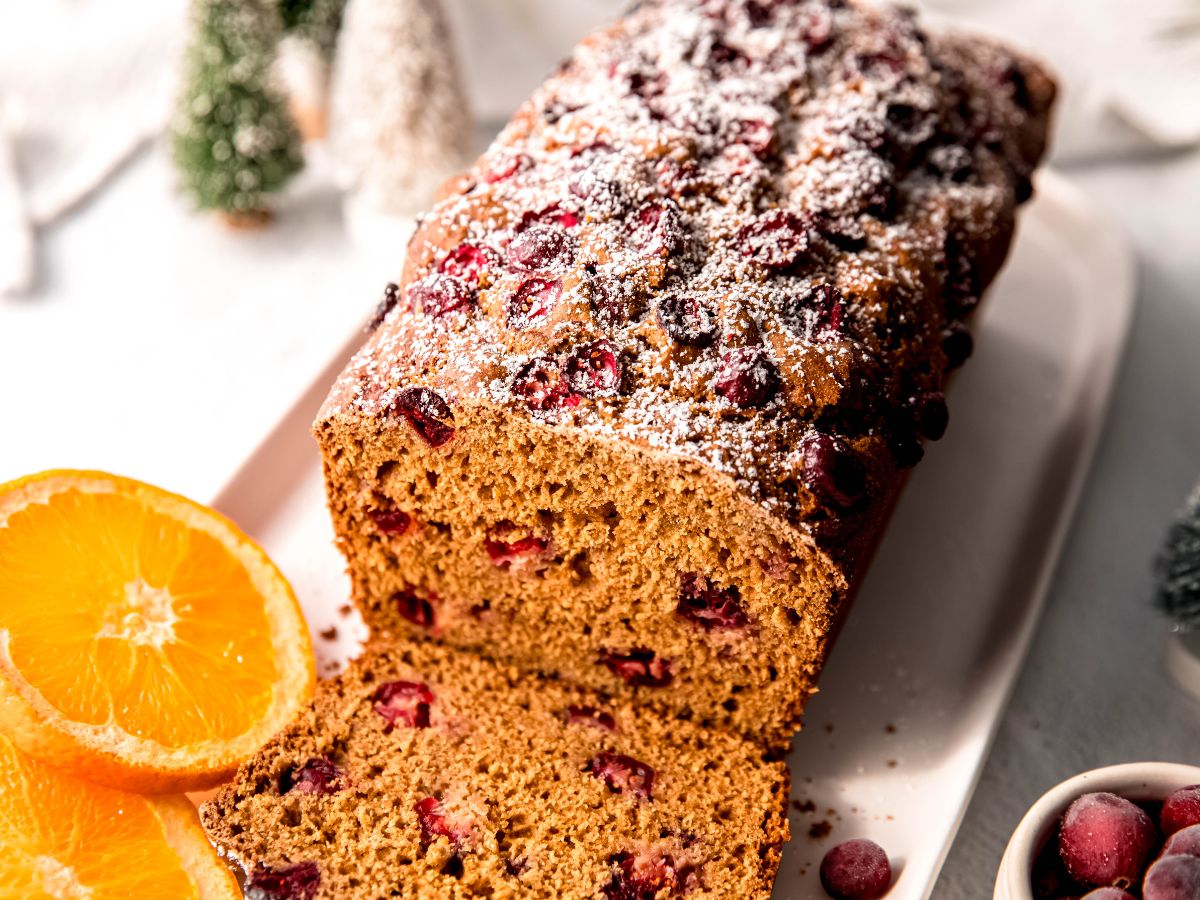 45 degree angle downward shot of a loaf of orange cranberry bread on a white serving platter with fresh orange slices and slightly frosted frozen cranberries.