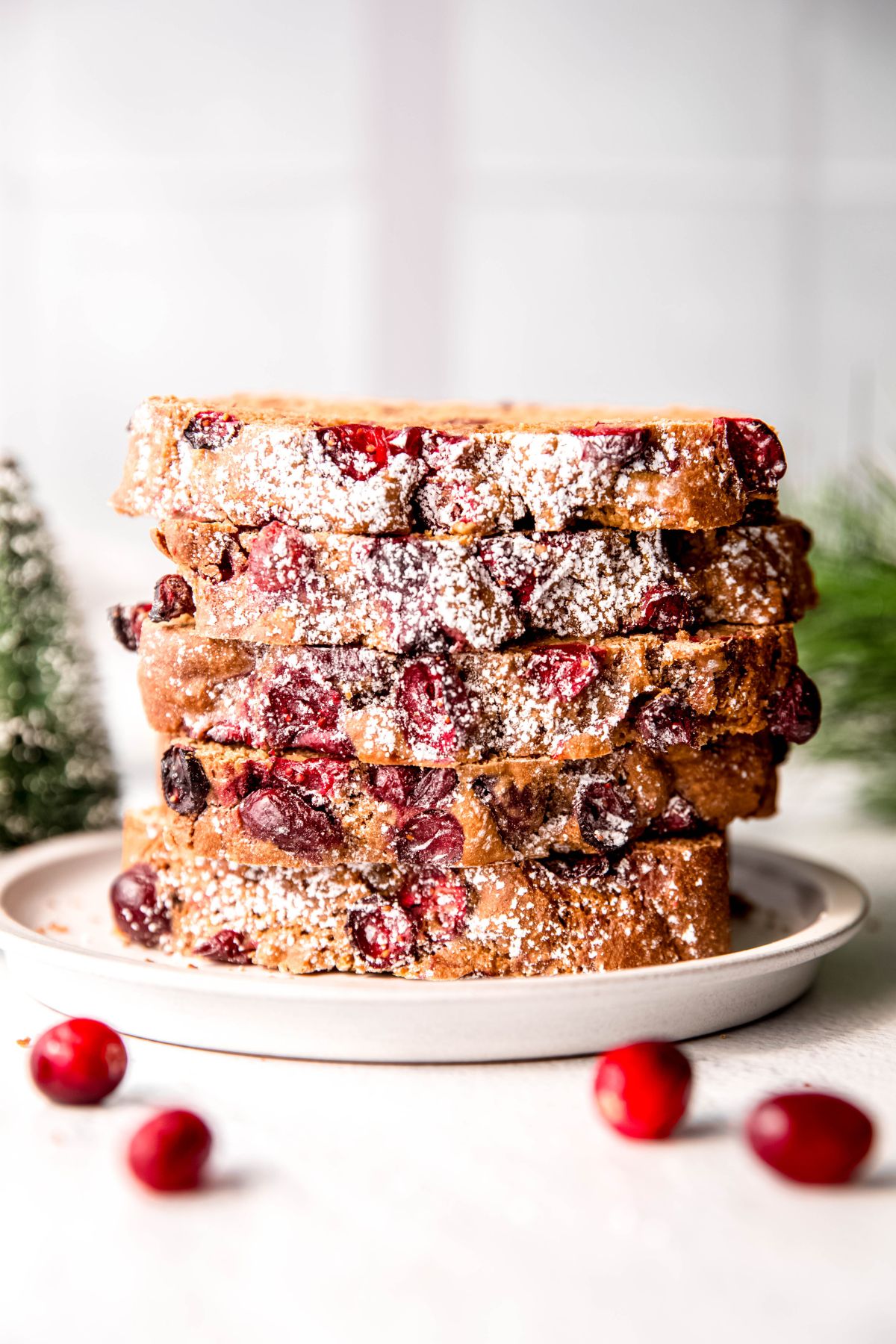 stack of 5 slices of homemade orange and cranberry bread on a white plate on a white table with fresh cranberries in the foreground and green pine tree sprigs in the background.