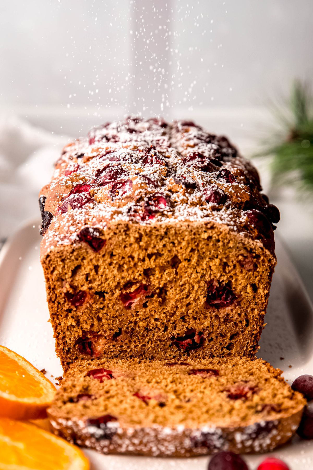 action shot of powdered sugar being sprinkled atop a loaf of whole wheat cranberry orange bread; it looks like snow falling.