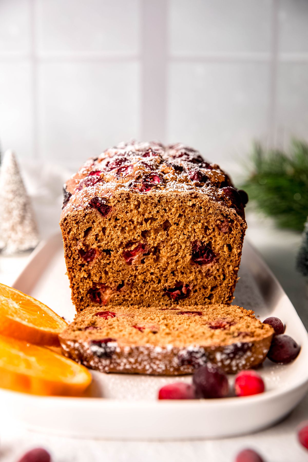 straight on view of a loaf of cranberry orange quick bread on a rectangular white plate on a white table; there's one slice that has its powdered sugar top facing the camera after being tilted forward.