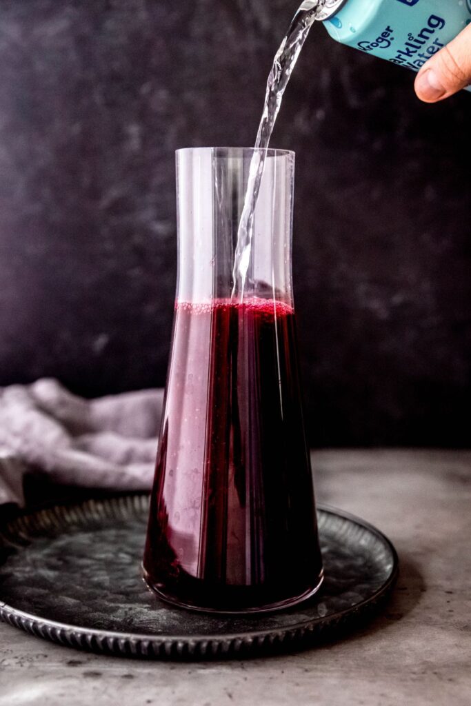 Pouring sparkling water into a large carafe of the blueberry pomegranate Halloween lemonade.