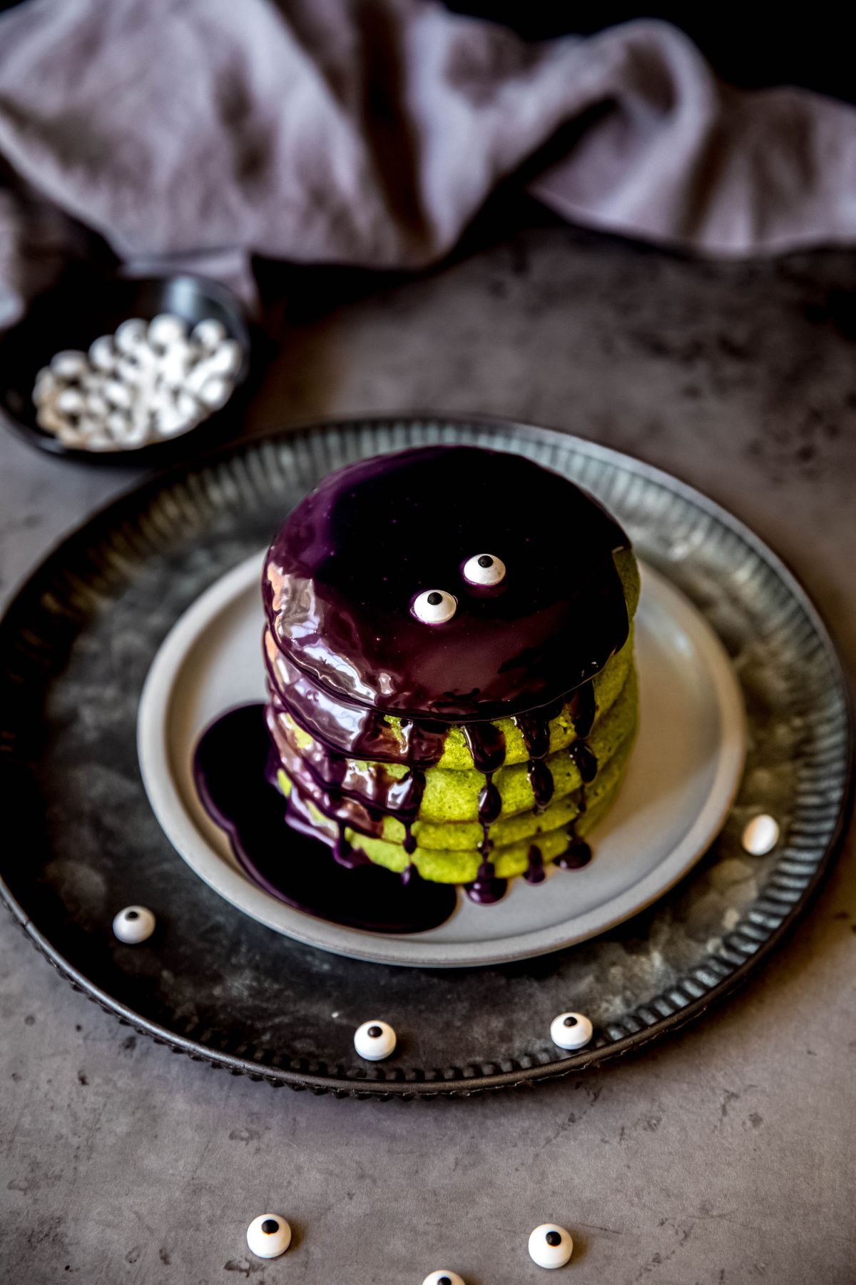 A overhead, angled shot of a stack of healthier halloween pancakes covered with ube pancake sauce and googly eyes.