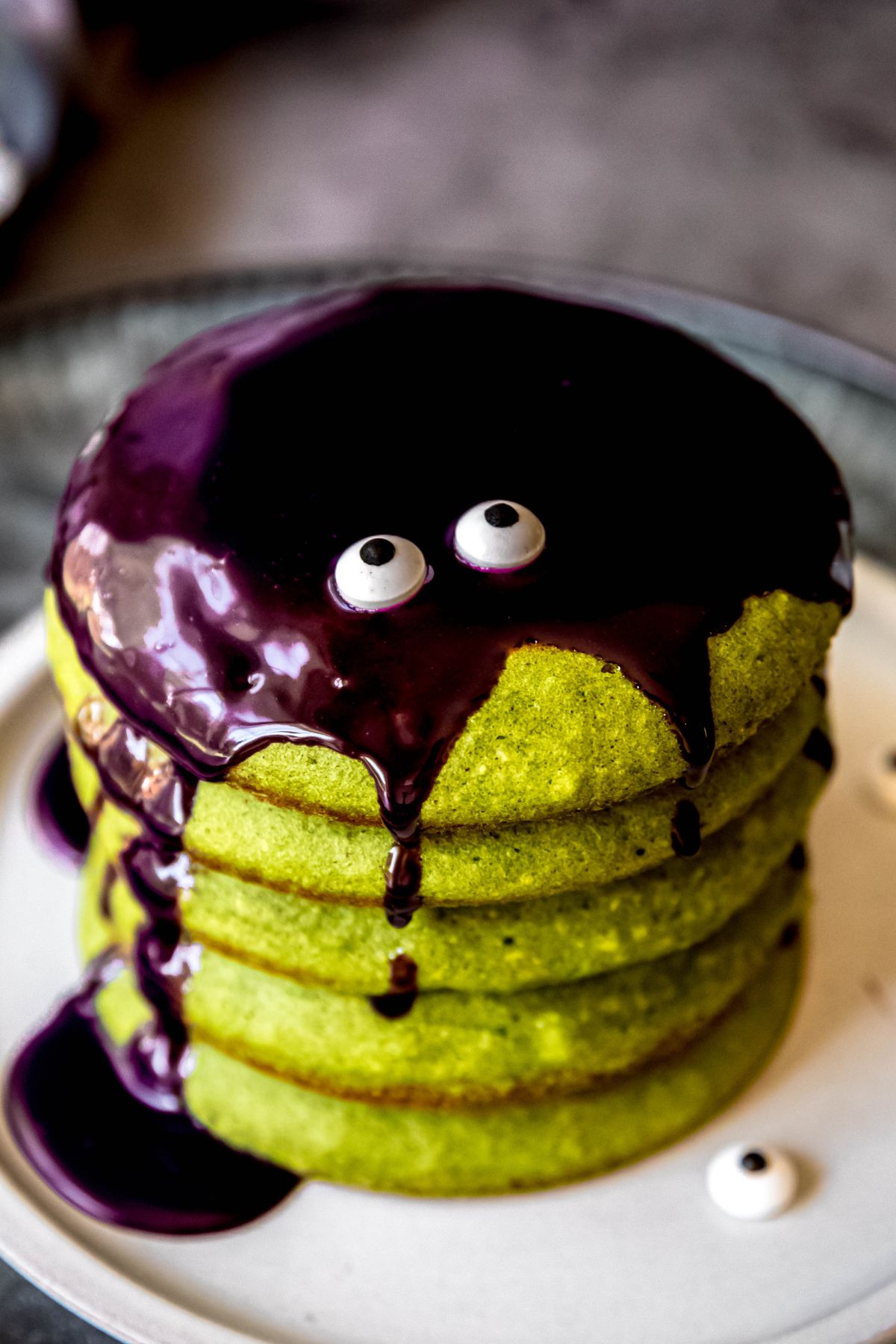 A close-up shot of a stack of green pancakes with purple syrup and googly eyes on top.