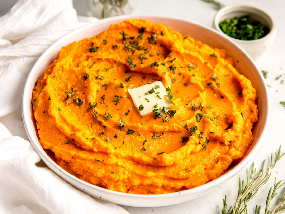 horizontal hero image of a bowl of savory garlic and herb mashed sweet potatoes on a white table with fresh herbs.