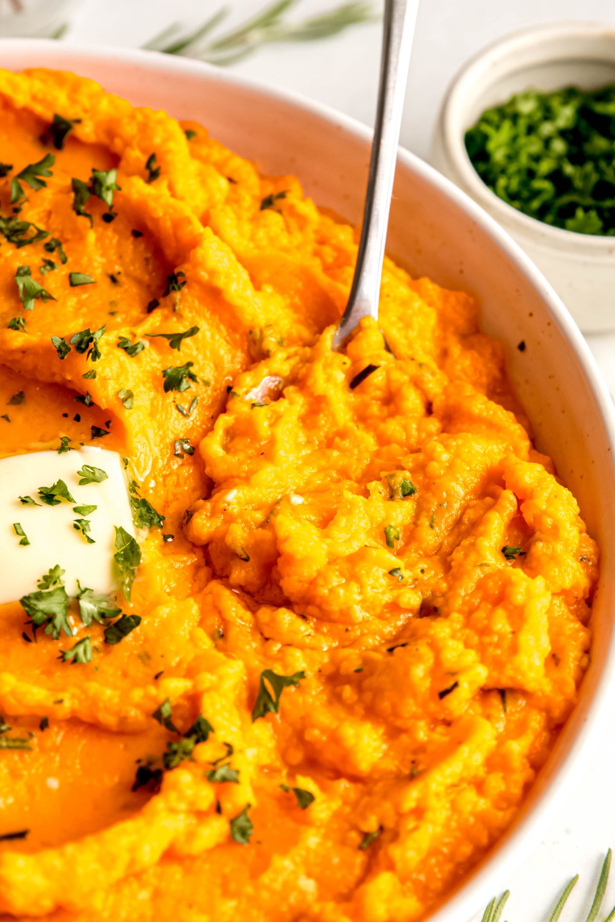 closeup shot of the right half of a white serving bowl filled with healthy mashed sweet potatoes with a silver spoon digging in to take a portion.