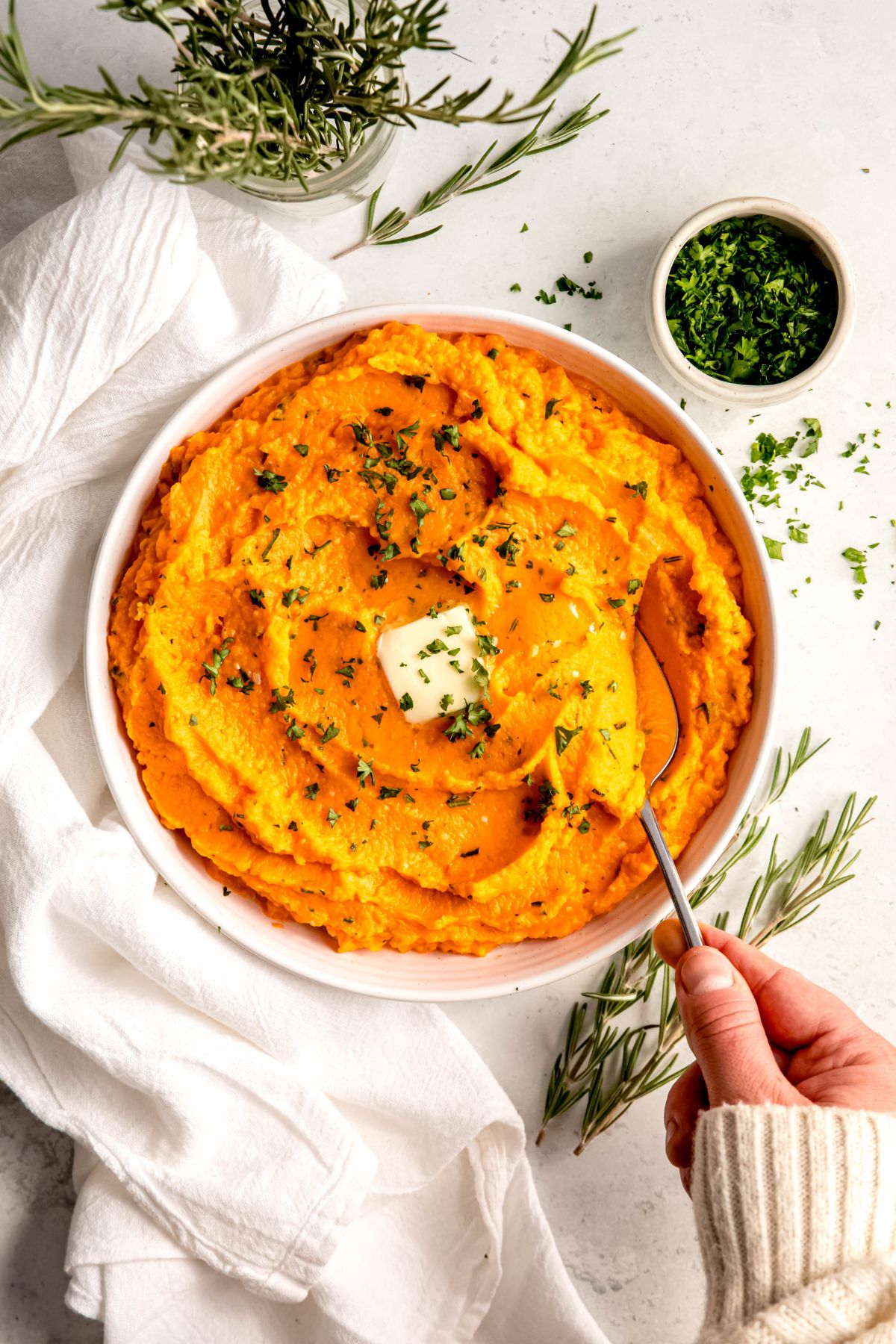 hand clad in a white sweater up to the knuckles using a silver serving spoon to scoop up a serving of herby garlic mashed sweet potatoes.