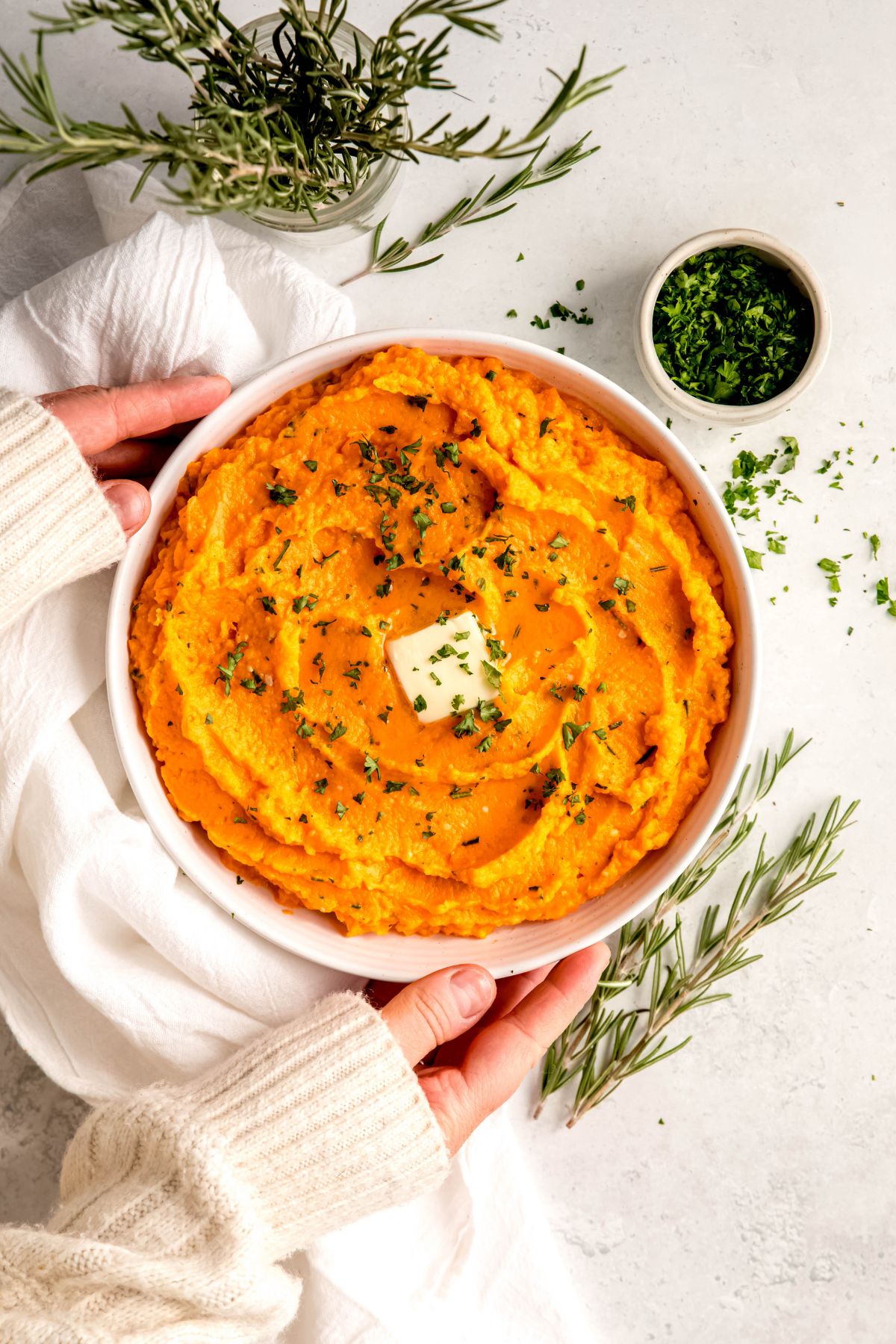 hands setting down a round white serving bowl filled with greek yogurt mashed sweet potatoes topped with a pad of butter and chopped herbs on a white table with sprigs of fresh rosemary in a jar and laying on the table.