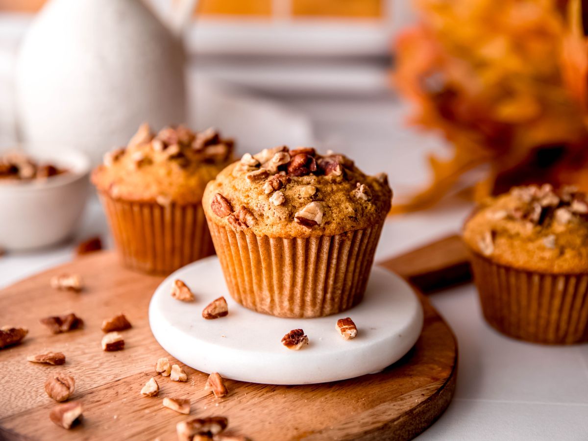 hero shot of 3 healthy whole wheat pecan pie muffins on a table with a bunch of orange leaves, more chopped pecans, and a white ceramic jug in the background in front of a orange sunset flowing through a white paned window.