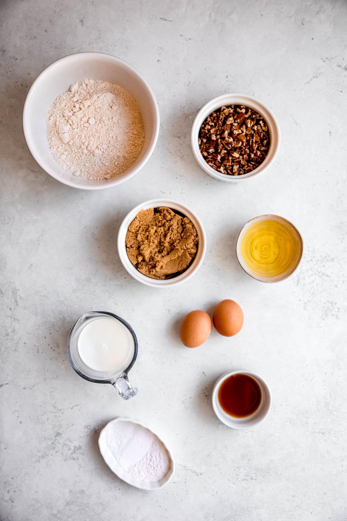 ingredients for making healthy pecan muffins measured out into white bowls on a white table.