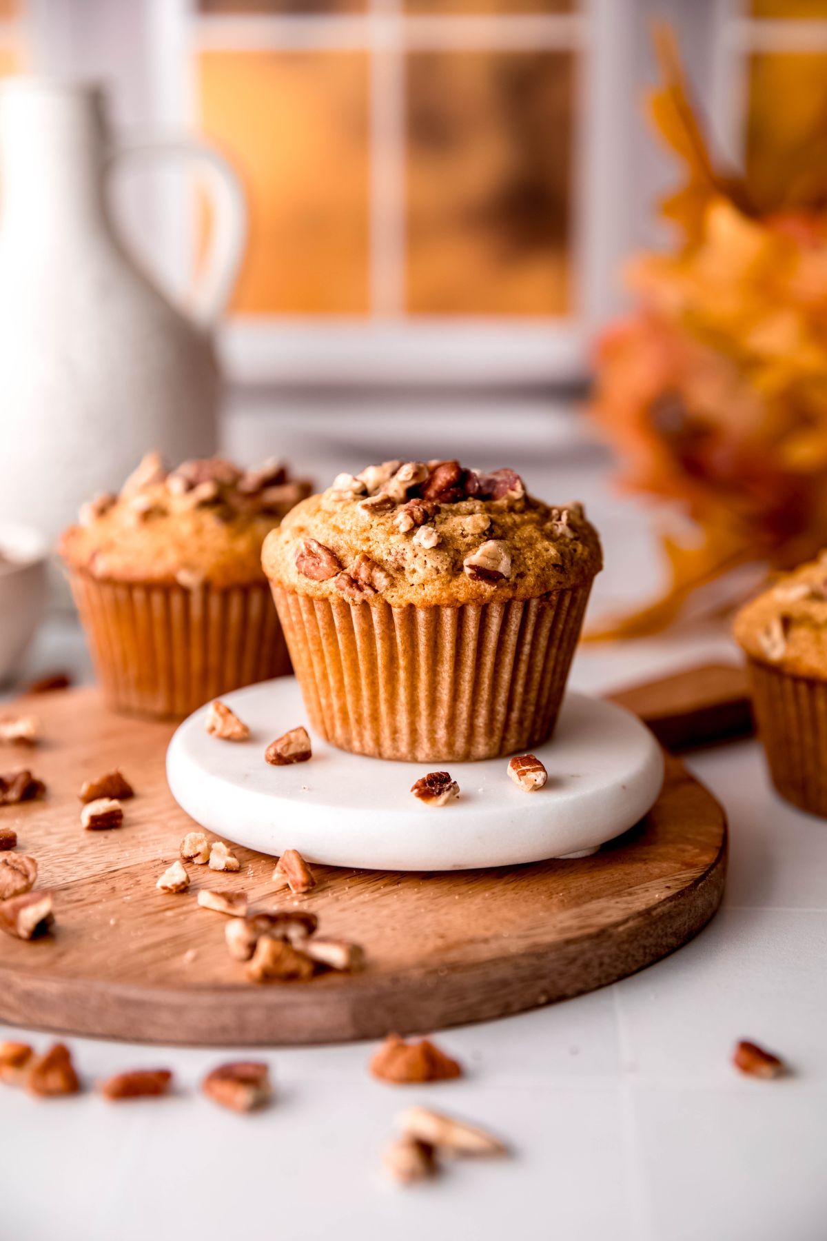three whole wheat pecan pie muffins on a table; the one in the middle is in focus while the other two are blurry; there's a warm window in the background with a sunset hue coming through.
