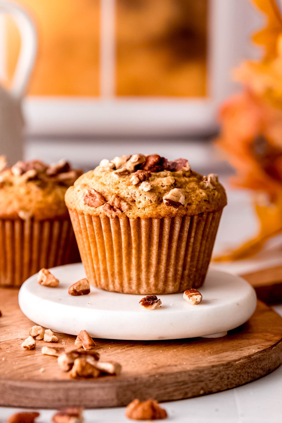 homemade pecan muffin on a white coaster on a wooden tray on a white tile table with chopped pecans scattered around and a warm fall-colored hue coming through the windows in the background.