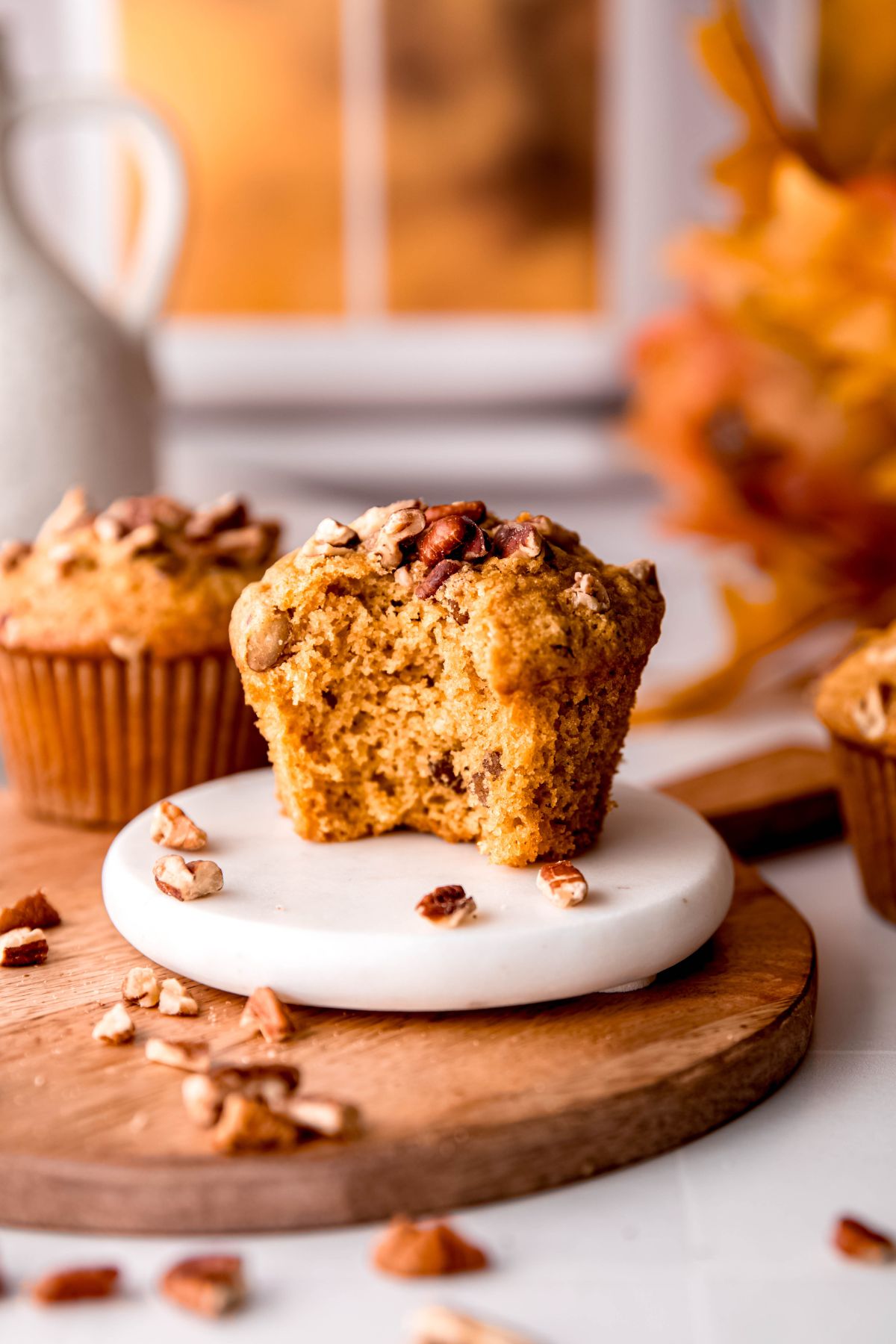 healthy pecan muffin with a bite taken out on a white coaster with a few pieces of pecans on top of a wooden tray with more pecans and a bunch of orange fall leaves blurred out in the background.