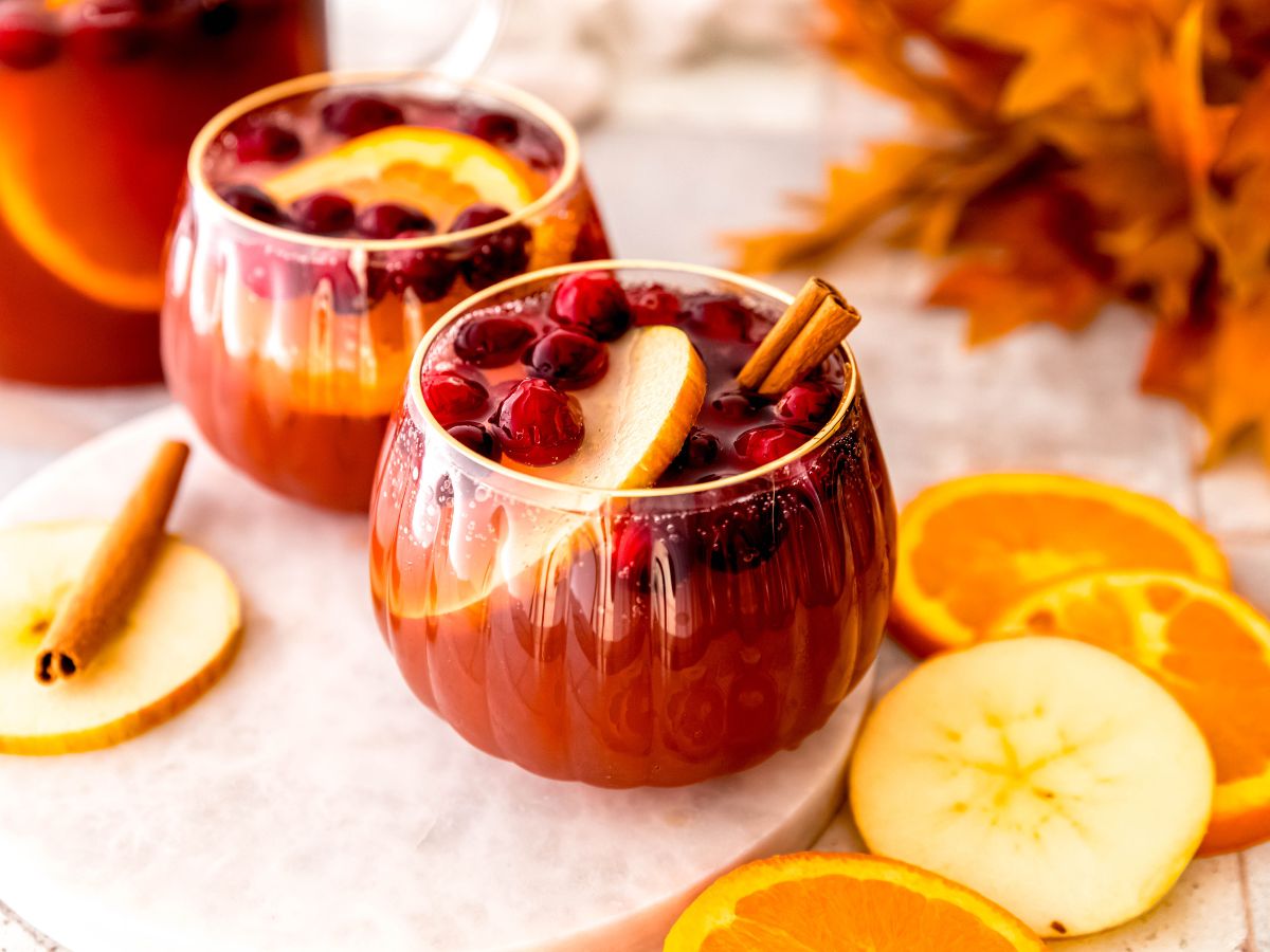 horizontal hero shot of two glasses of non-alcoholic thanksgiving punch on a white table with a bunch of orange fall leaves, slices of orange and apple, and a pitcher with the rest of the punch.
