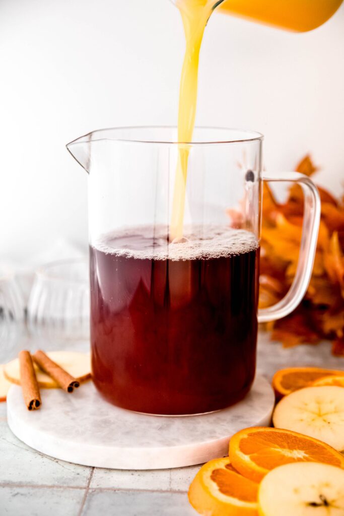 orange juice being poured into the pitcher with the apple cider, pomegranate juice, and simple syrup.
