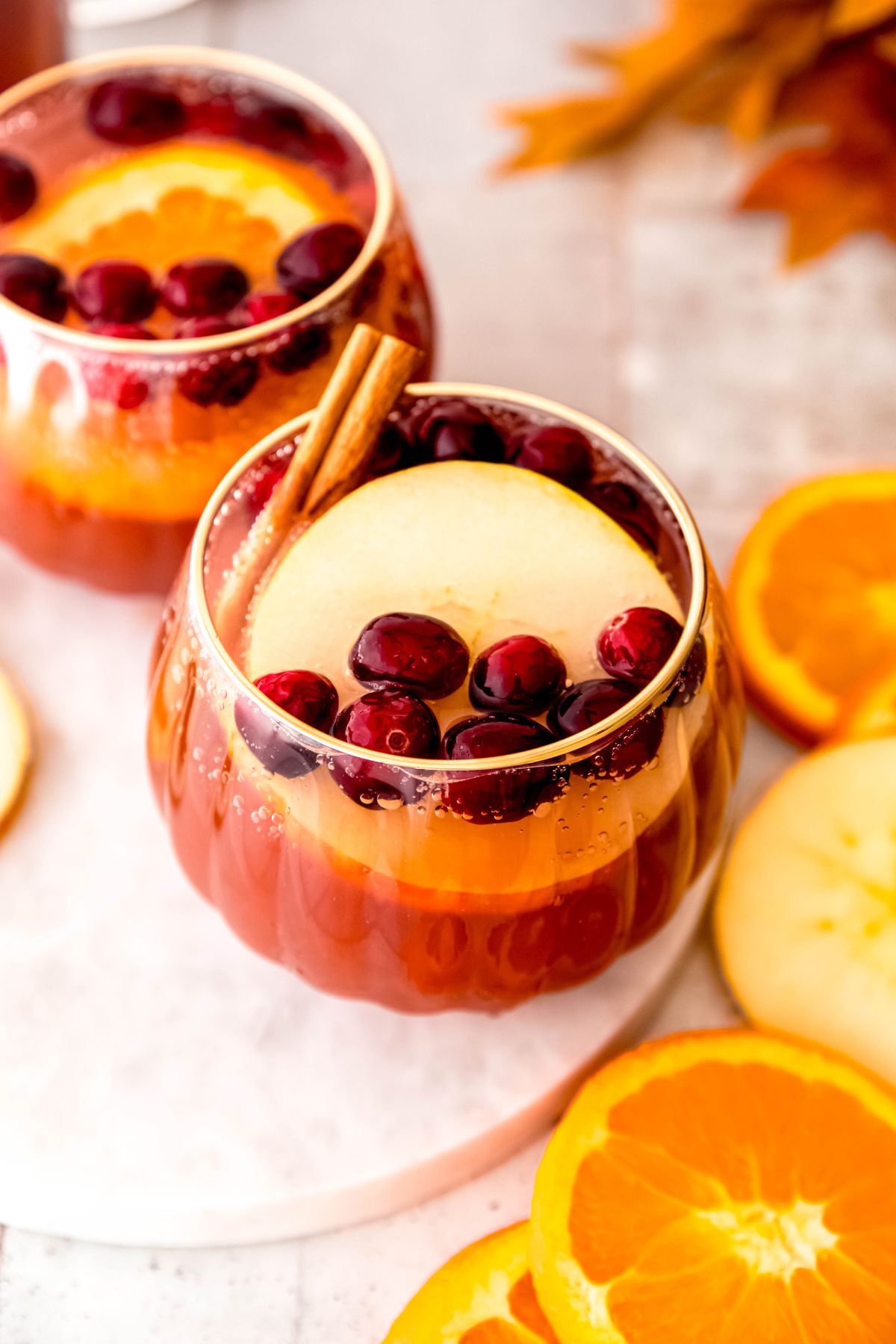 two clear scalloped punch glasses filled with non alcoholic Thanksgiving punch on a white table with slices of orange and apple on the table.