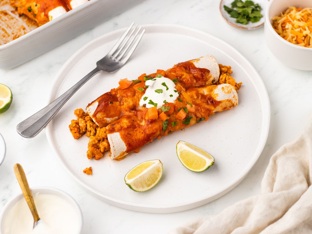 white dinner plate with two turkey enchiladas topped with crema and cilantro with two lime wedges on the side on a white table with a bowl of extra crema, a pinch bowl of cilantro, a bowl of shredded cheese, and the rest of the casserole dish surrounding it.
