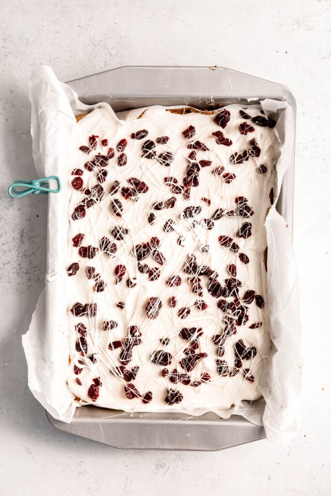 cranberry bliss bars in the pan after adding the cream cheese frosting, dried cranberries, and white chocolate drizzle.