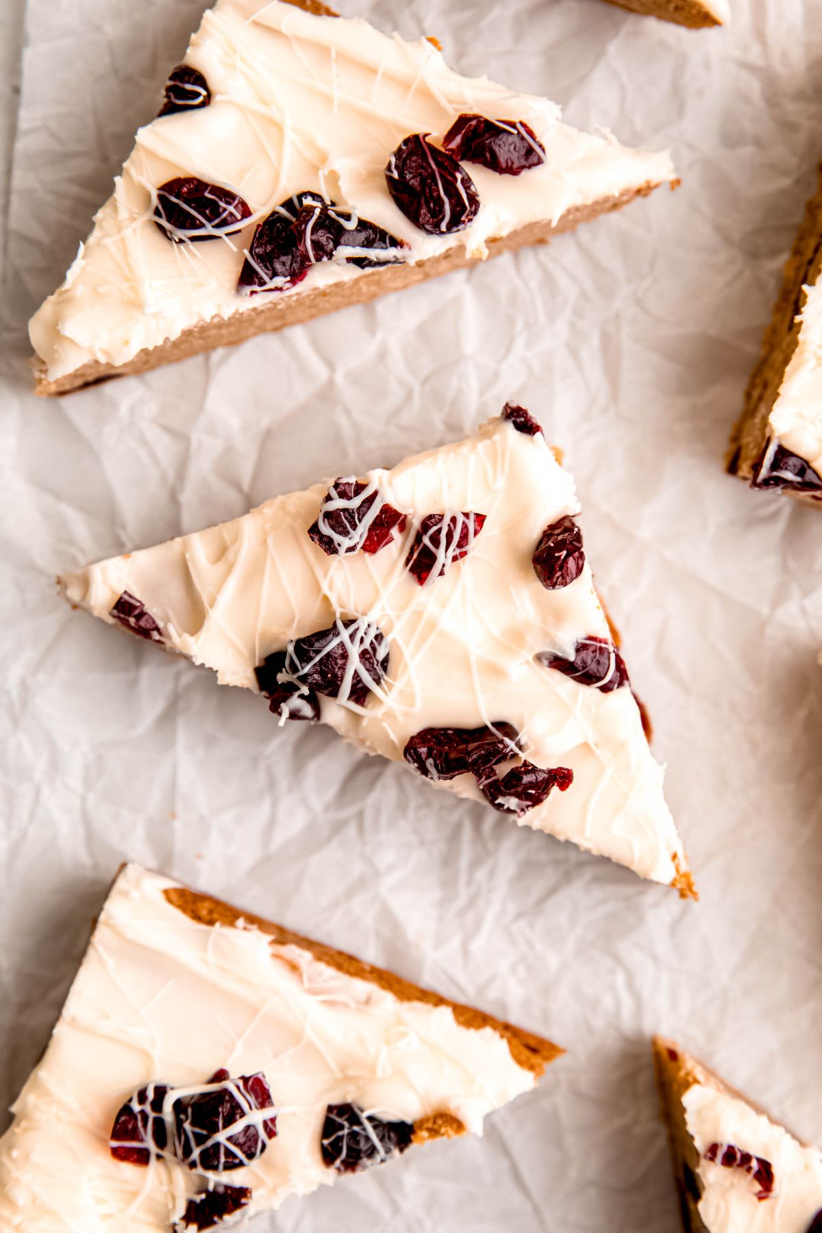 overhead shot of a homemade cranberry bliss bars on a piece of crumpled white parchment paper showing the pretty tops coated in cream cheese frosting, dark red dried cranberries, and a white chocolate drizzle.