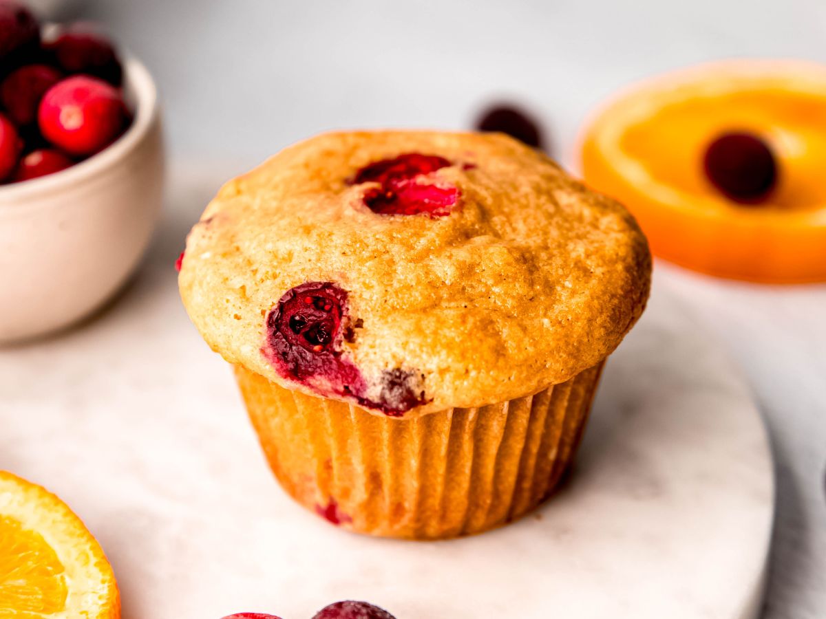 horizontal hero shot of a cranberry orange protein muffin on a white table with a small white bowl of fresh cranberries to the back left and a round slice of fresh orange topped with a fresh cranberry on the right; only the muffin is in focus.
