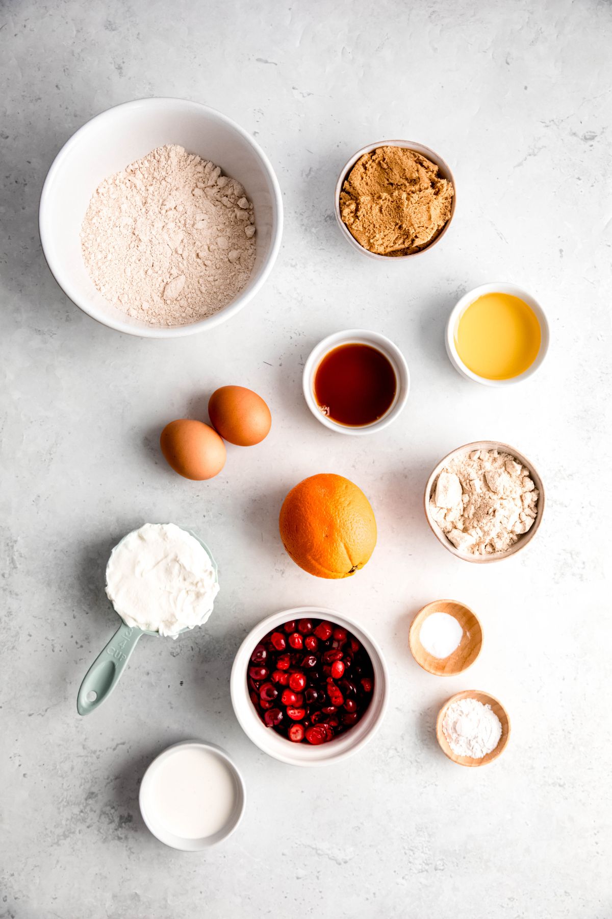 overhead shot of the ingredients needed to make cranberry orange muffins with protein powder measured out in bowls on a white table.