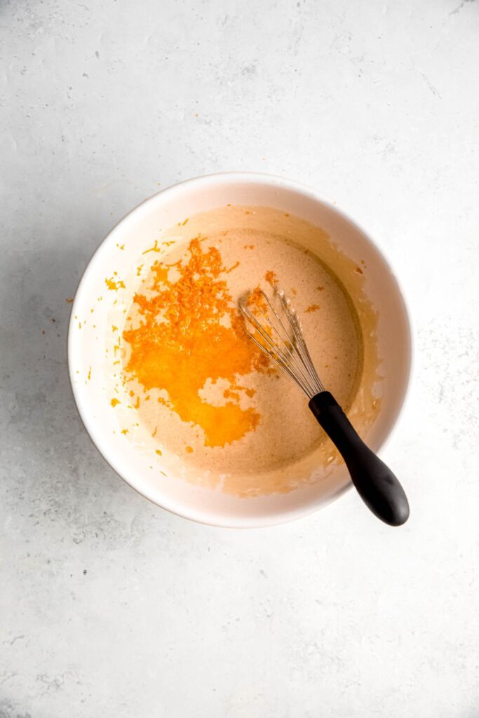 orange zest, orange juice, and oil added to the bowl with the wet ingredients; there is a black-handled whisk in the bowl.