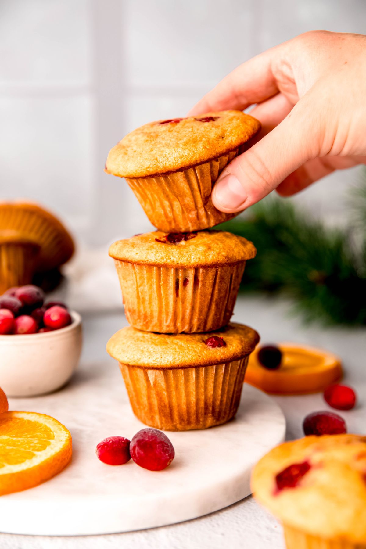 hand grabbing the top of a stack of 3 cranberry orange-flavored protein muffins on a white table.