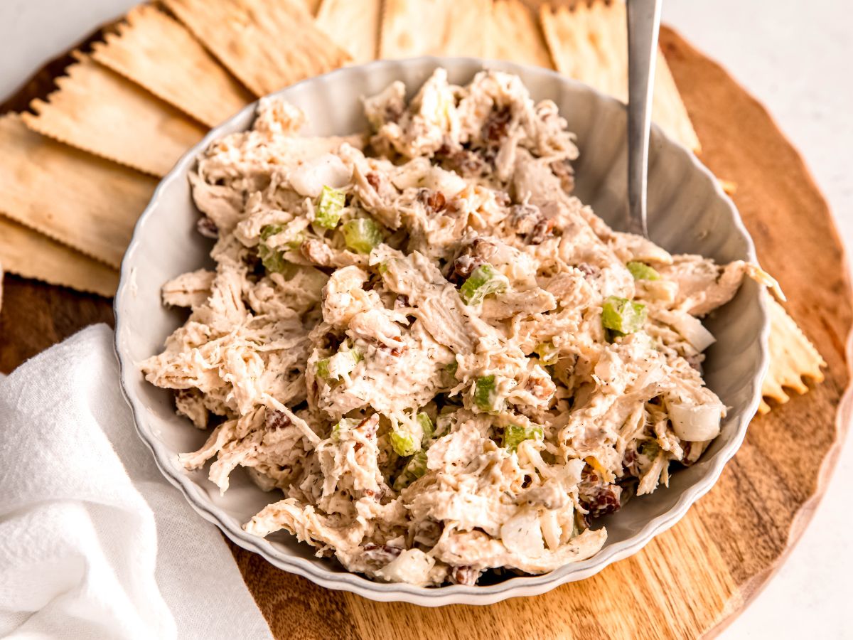 horizontal hero shot of a snacking board with a white bowl of turkey salad sandwich filling with a silver spoon surrounded by thin crackers.