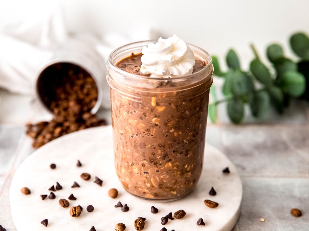 horizontally framed hero shot of a mason jar of mocha protein overnight oats on a white marble round atop a grey slate table with mini chocolate chips and espresso beans scattered around and a green houseplant peeking into the frame blurred out in the background.