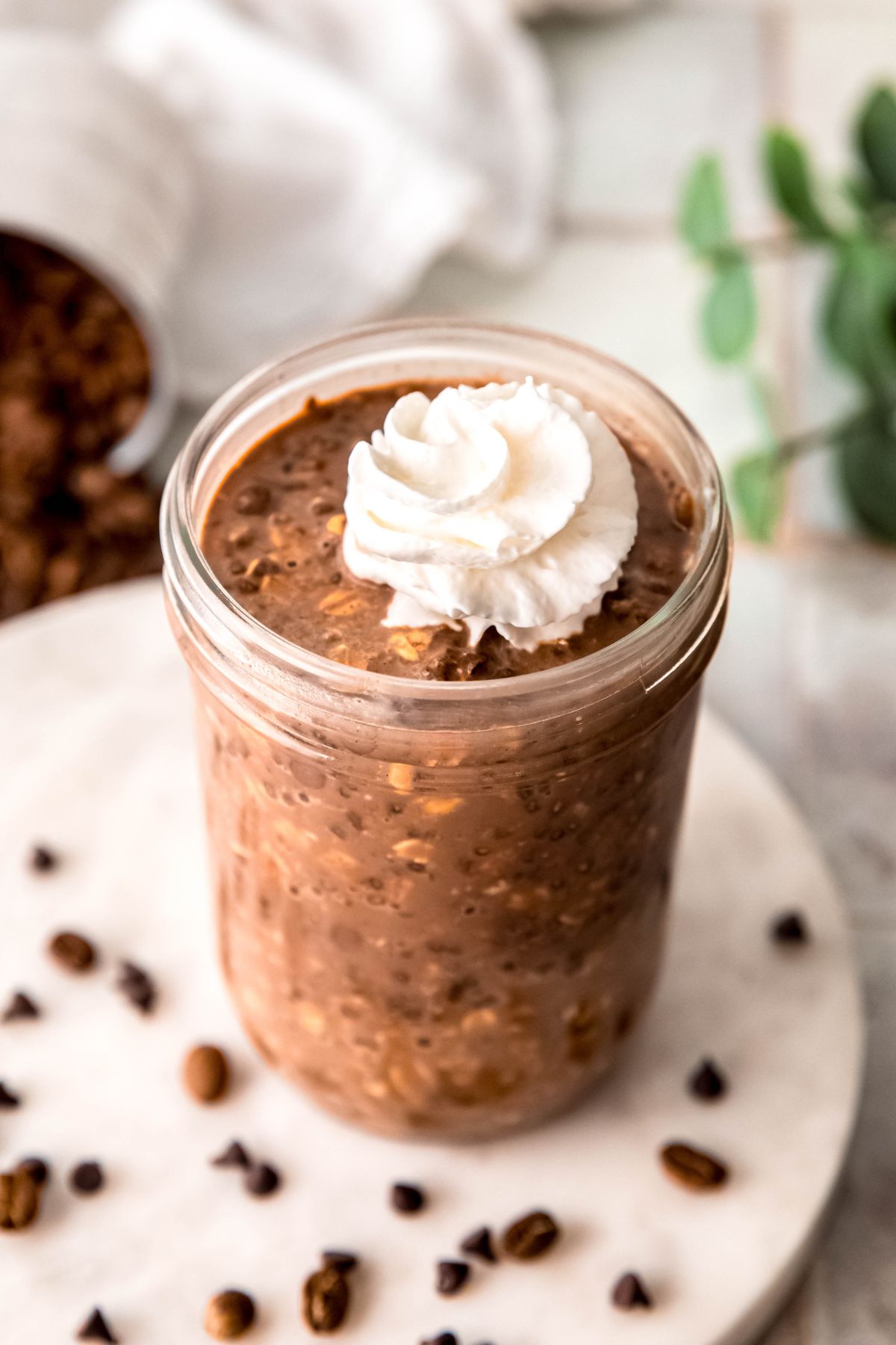 closeup 45 degree angle shot of a jar of mocha flavored high protein overnight oats with a squirt of whipped cream as garnish with the white table, espresso beans, and mini chocolate chips blurred out below.