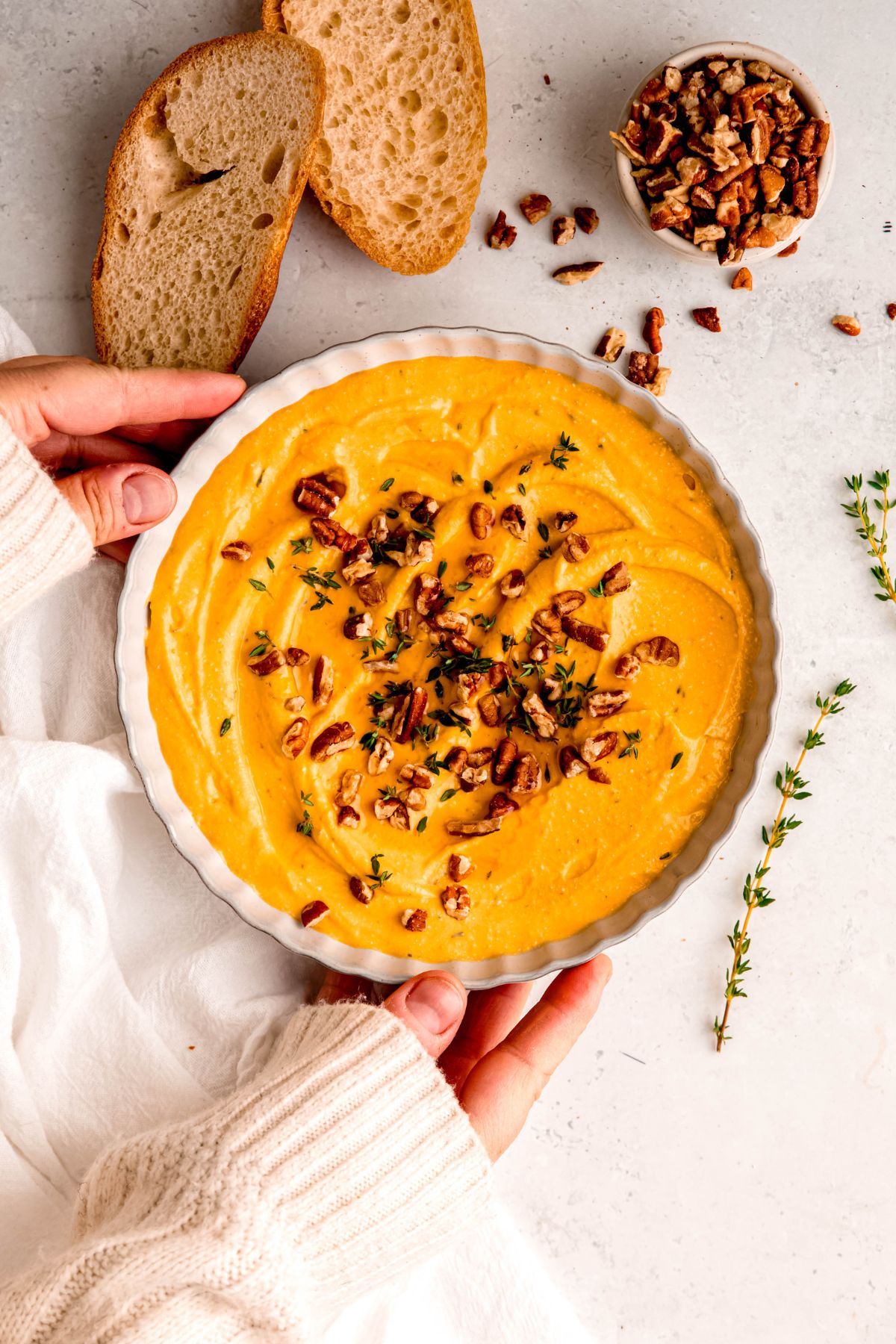 Two hands holding a bowl of pumpkin whipped feta next to a small cup of nuts and toasted baguette slices.