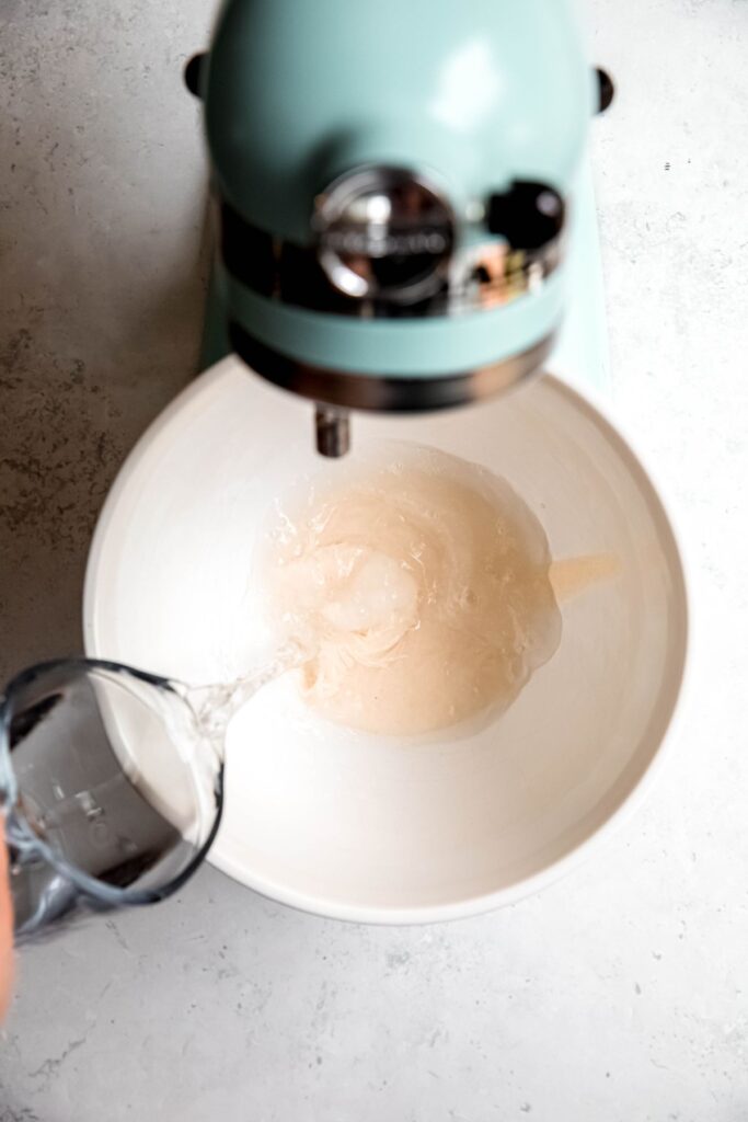 action shot of water being poured into a stand mixer's bowl with the sourdough discard, sugar and yeast.
