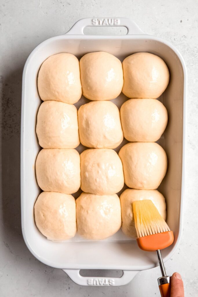 action shot of a hand using a pastry brush to brush the tops of the fully proofed rolls with egg wash; the rolls are all snug and touching.