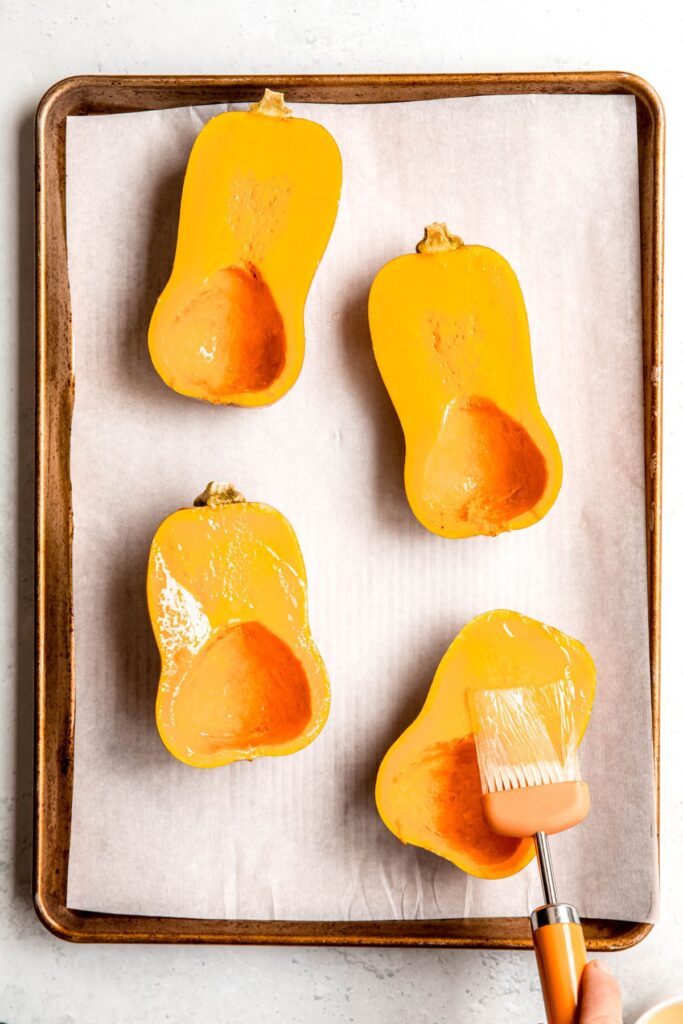 two halved honeynut squash placed cut side up on a parchment lined baking sheet with a hand actively brushing the tops with avocado oil.