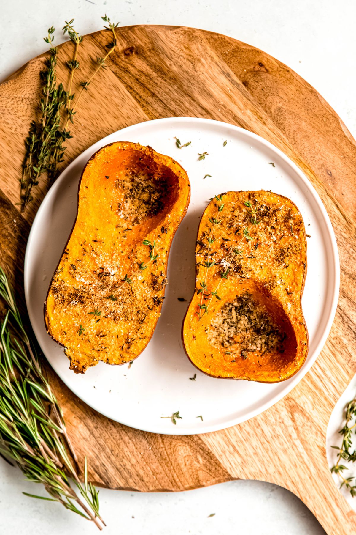 round white plate with two halves of parmesan and herb roasted honeynut squash garnished with a few fresh sprigs of thyme on a blonde wooden surface with more fresh thyme and fresh rosemary on the board.