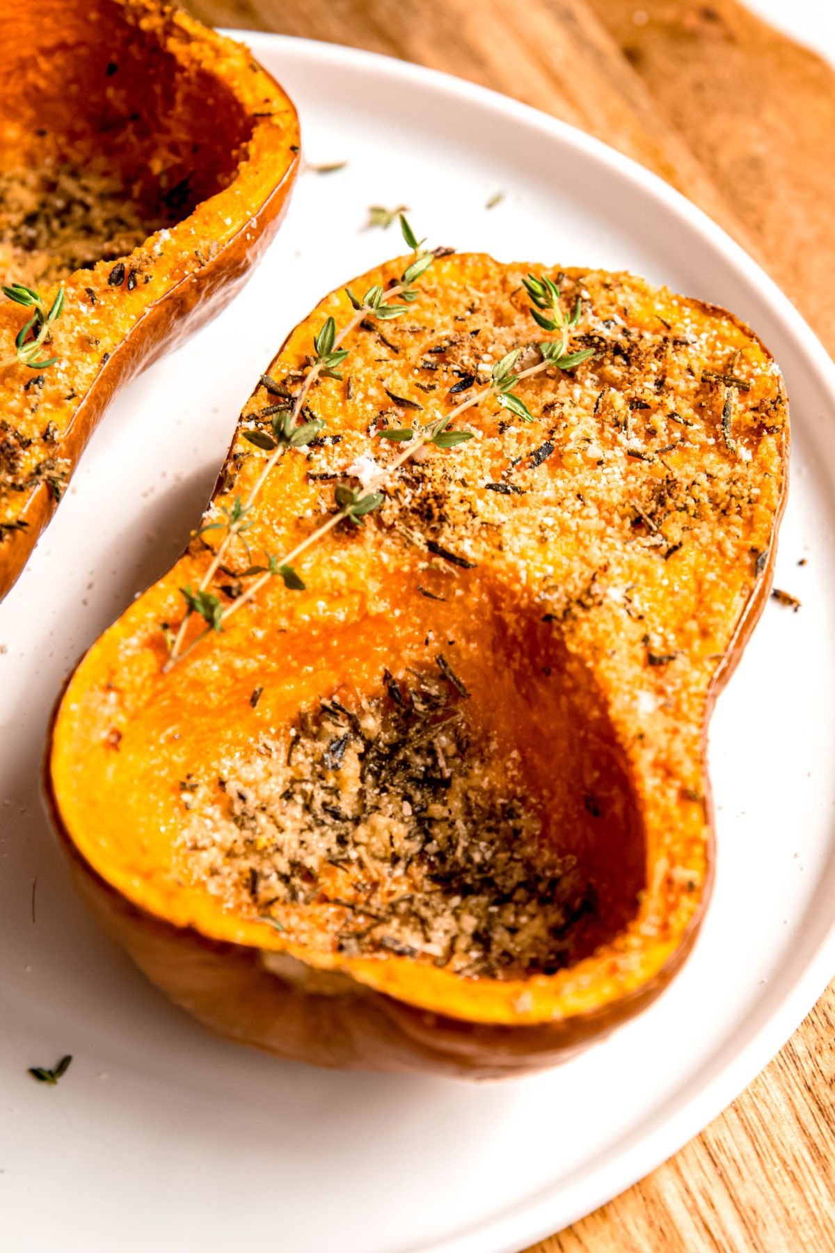 45 degree angle closeup shot of a half of a roasted honeynut squash topped with parmesan, rosemary, and thyme; the small bit of the skin you can see on the bottom half is slightly wrinkled and golden brown.