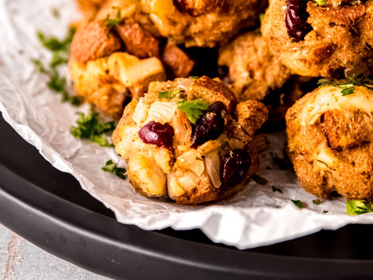 horizontal hero image of a closeup shot of a plate of baked cranberry turkey stuffing balls garnished with parsley on a piece of white parchment paper on a black plate.