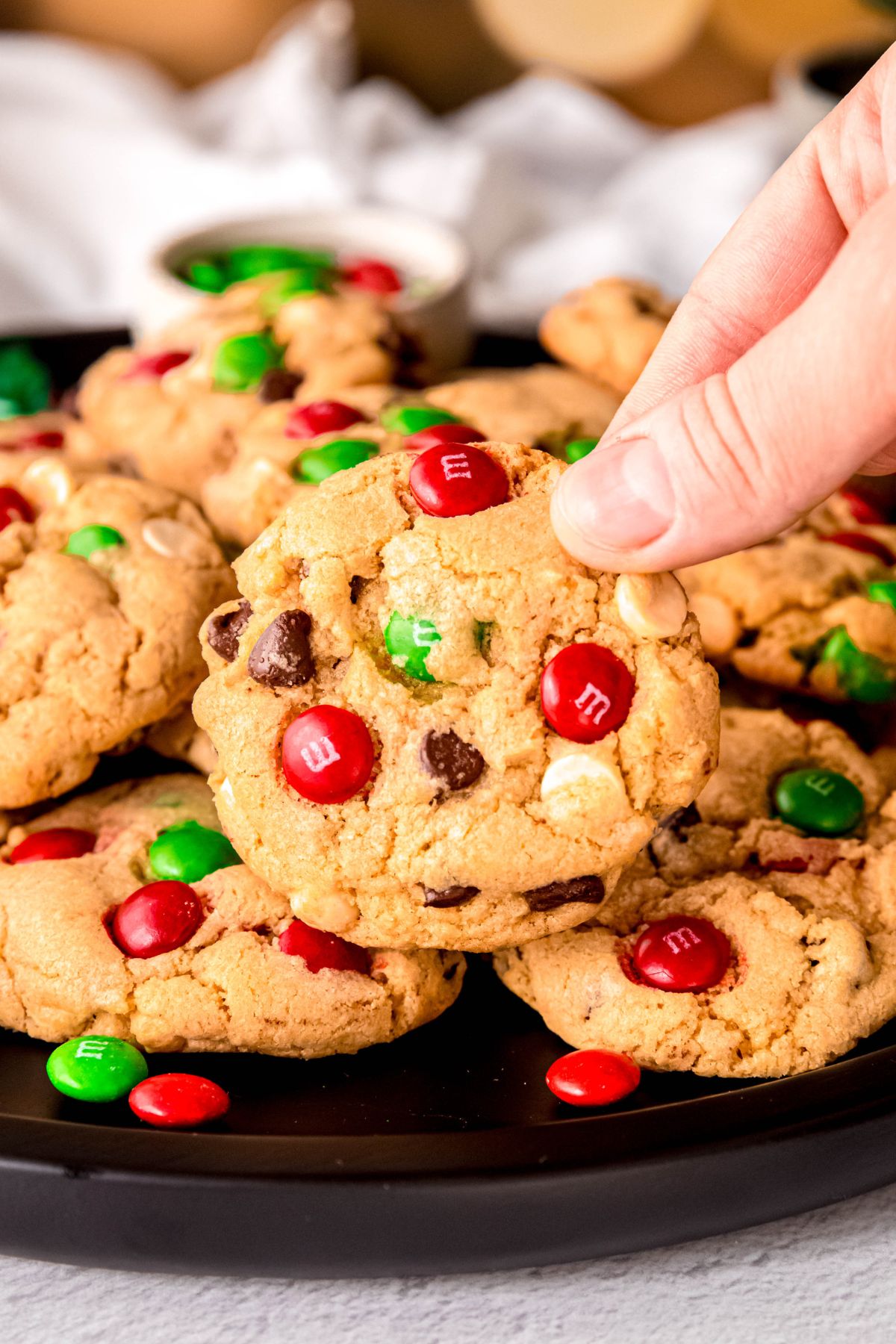 hand grabbing a Christmas themed loaded chocolate chip cookie off the top of a pile of the cookies on a black plate.