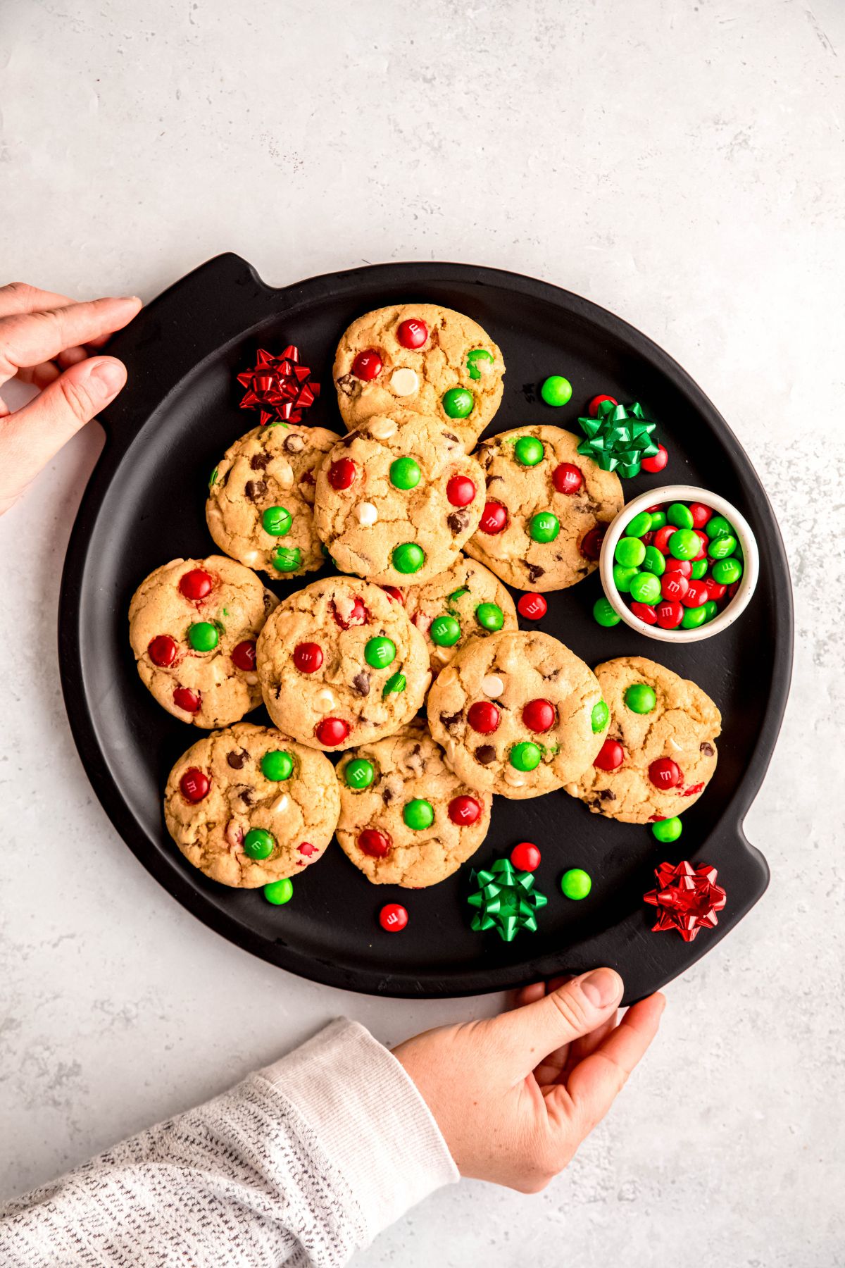 hands in a white sweater setting down a black serving tray of Christmas chocolate chip cookies decorated with extra green and red M&Ms and a few small red and green bows.