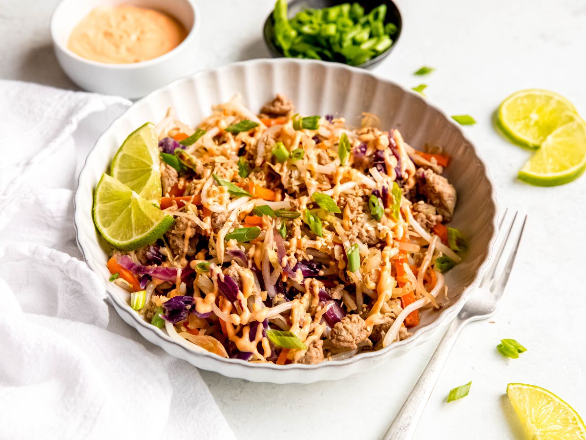 A horizontal shot of a large bowl of egg roll in a bowl on the counter with limes, green onions, and spicy mayo.
