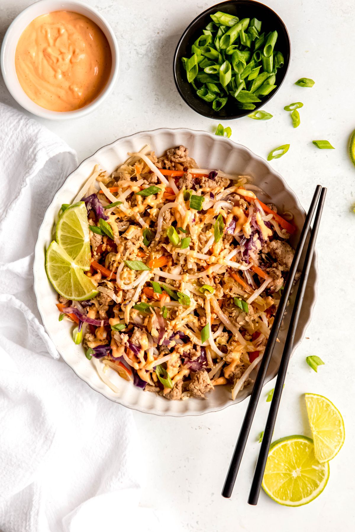 An overhead shot of a serving of egg roll in a bowl drizzled with spicy mayo with black chopsticks, limes, green onions, and spicy mayo.
