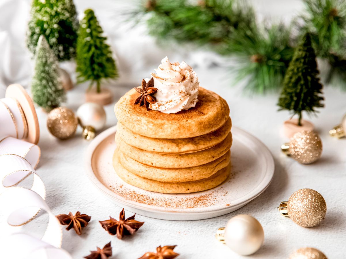 horizontal hero shot of a plate of whipped cream-garnished eggnog pancakes on a white table with green bottle brush trees, a sprig of fresh pine, gold and white mini ornaments, whole star anise pods, and a partially unwound roll of gold-edged white ribbon on a wooden spool.