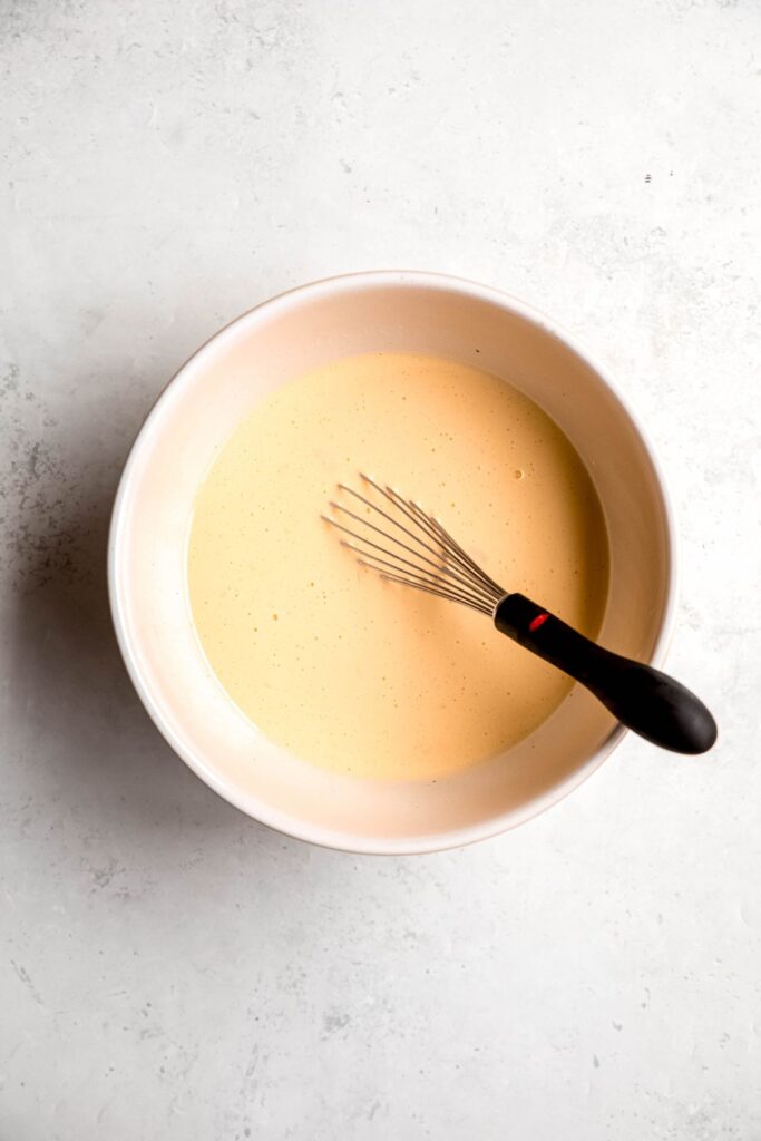 white mixing bowl with whisked wet ingredients and a black-handled whisk.