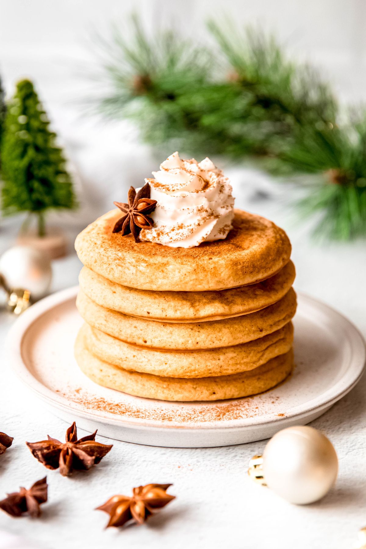 stack of fluffy eggnog pancakes on a white plate on a white table with whole star anise, white christmas ornaments, and green sprigs of pine; the stack is topped with whipped cream and a sprinkle of cinnamon.
