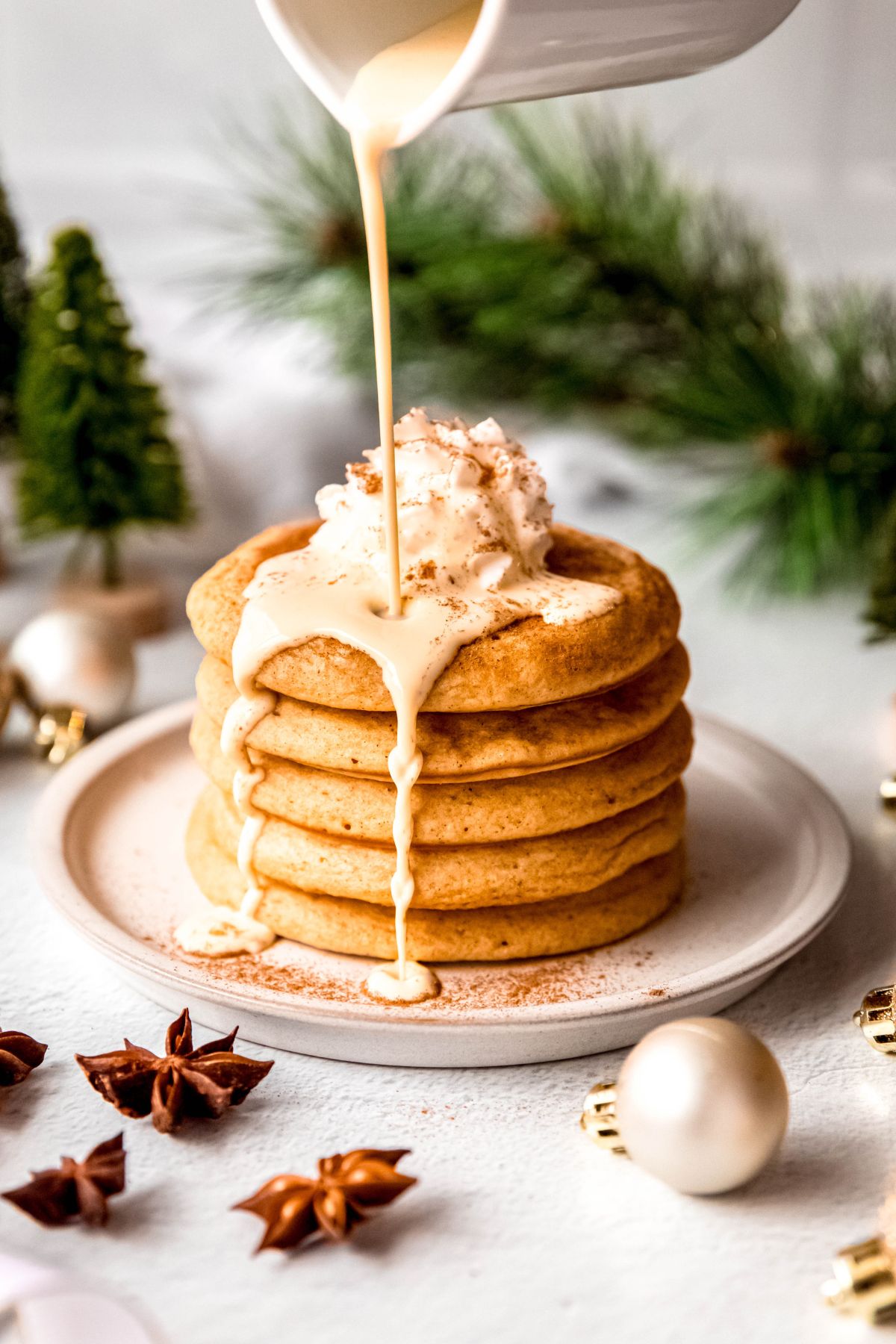 action shot of eggnog being poured on a fluffy stack of eggnog pancakes topped with whipped cream and cinnamon.