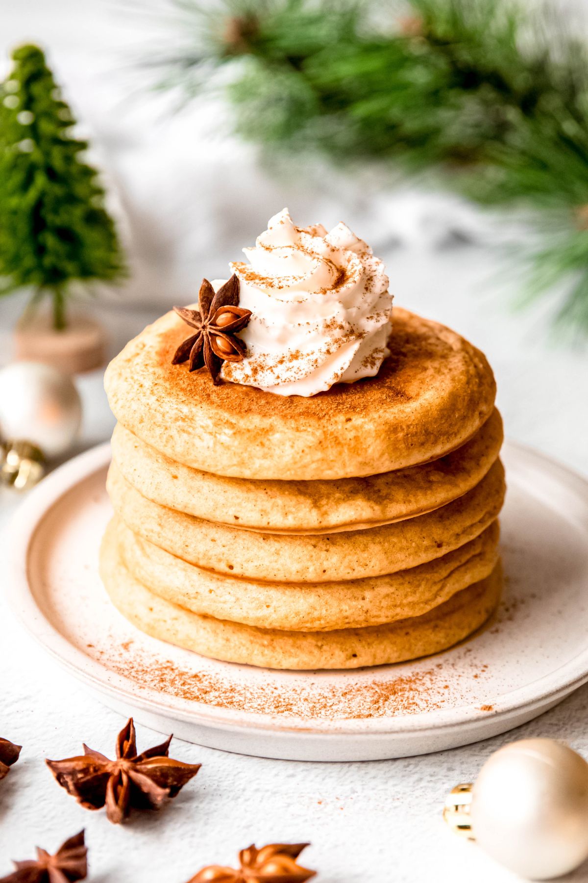 eggnog christmas pancakes in a stack on a white plate on a white table with whole star anise pods, matte white christmas ornaments, green bottle brush trees, and a sprig of pine framing the shot.
