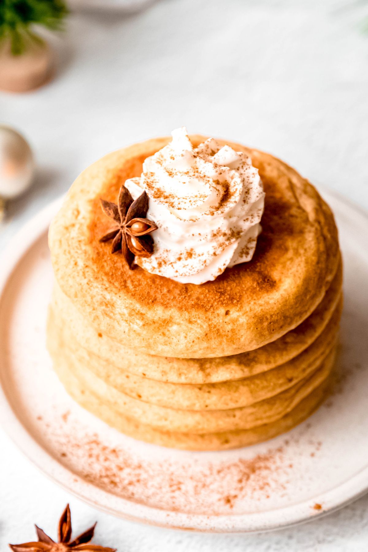 closeup 45 degree angle shot of a stack of eggnog whole wheat pancakes topped with whipped cream, cinnamon, and a whole star anise on a white plate on a white table.