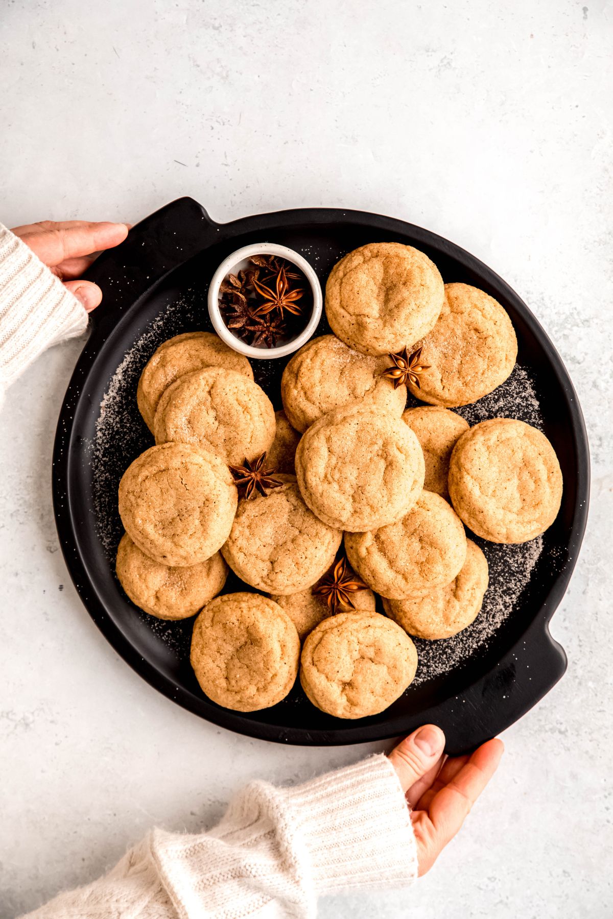 hands in a white sweater up to the knuckles setting down a black serving tray of homemade eggnog snickerdoodle cookies on a white table