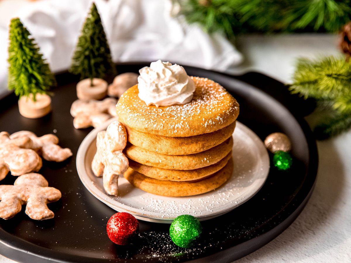 horizontally framed hero shot of gingerbread pancakes served in a stack with powdered sugar, whipped cream, and a mini gingerbread man leaning against the stack on a wintry table with bottle brush trees and christmas ornaments.