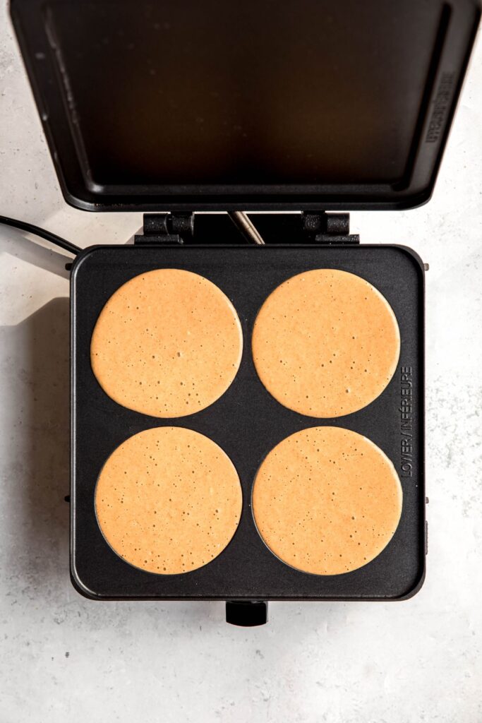 4 rounds of raw pancake batter on a pancake griddle with tiny bubbles starting to form on top.