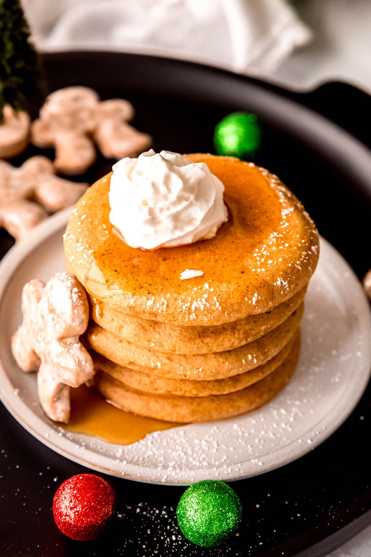 stack of healthy gingerbread pancakes topped with whipped cream, maple syrup, and powdered sugar on a plate with a mini gingerbread man.